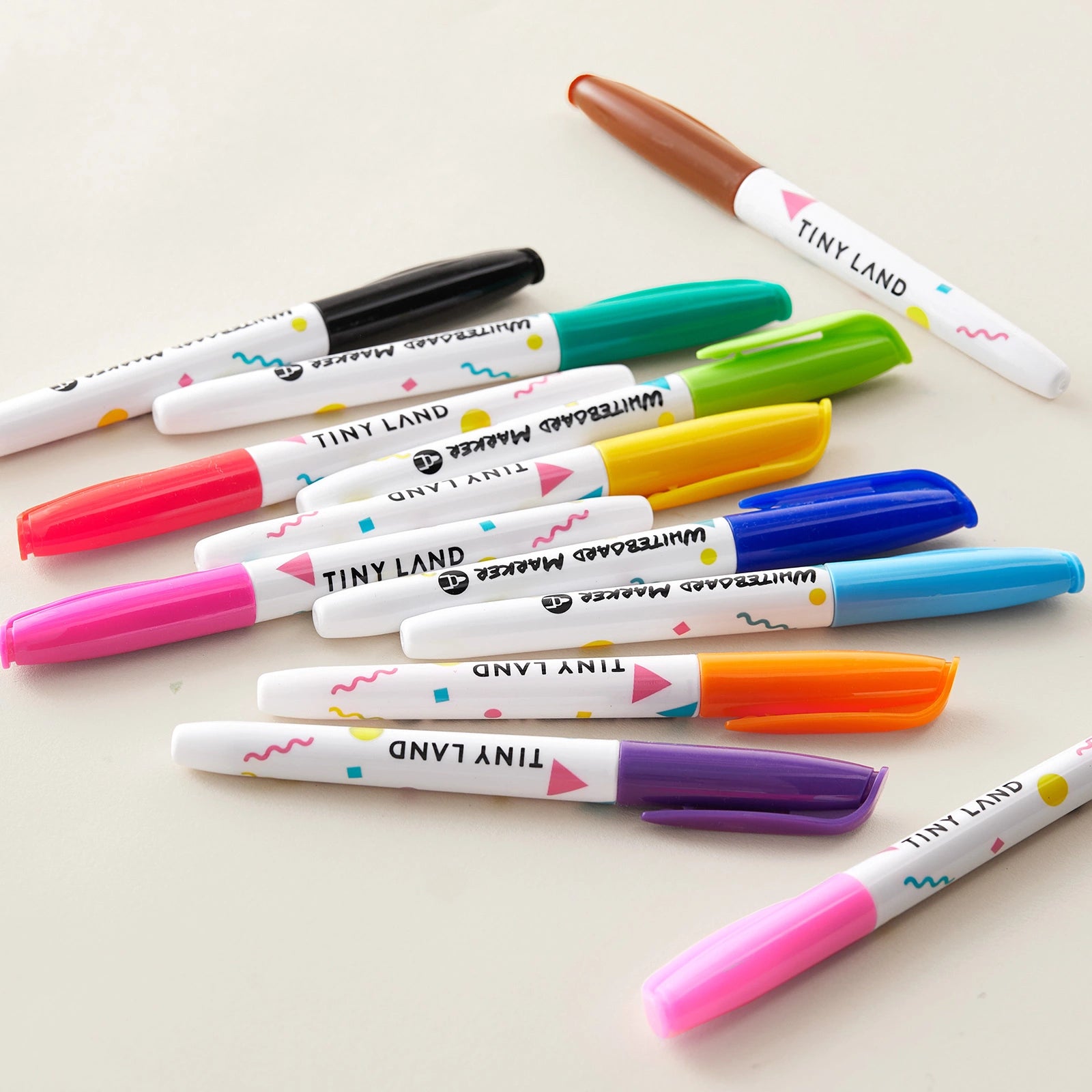 A pile of colorful marker pens with patterned white barrels from the Tiny Land® Magnitales Easel Art Kit features blue, pink, purple, orange, and brown caps. The Tiny Land brand is printed on each pen.