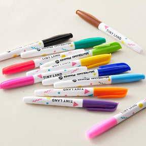 A pile of colorful marker pens with patterned white barrels from the Tiny Land® Magnitales Easel Art Kit features blue, pink, purple, orange, and brown caps. The Tiny Land brand is printed on each pen.