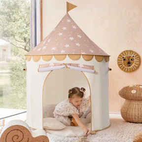 A young child plays inside the Tiny Land® Cheerful Pennant Castle Tent, a cream-colored kids' playhouse with a star-patterned roof and flag, set on a soft rug near wicker baskets and a wall featuring a cheerful lion face.