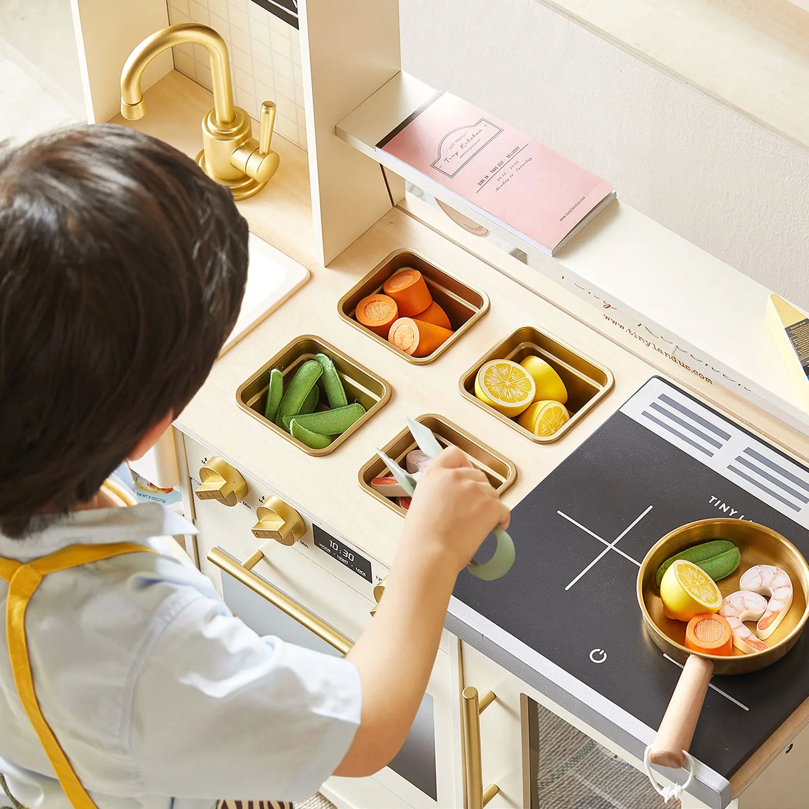 A child in an apron enjoys pretend play with the Tiny Land® BistroSwitch Play Kitchen, using tongs to pick up toy vegetables. The Tiny Land set features a stove, sink, compartments, and play food like carrots, peas, lemon slices, and shrimp.|White