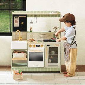A young child dressed as a chef enjoys pretend play with the Tiny Land® BistroSwitch Play Kitchen, featuring a sink, fridge, oven, stovetop, shelves, and fun toy utensils and food from the Tiny Land brand.|Green