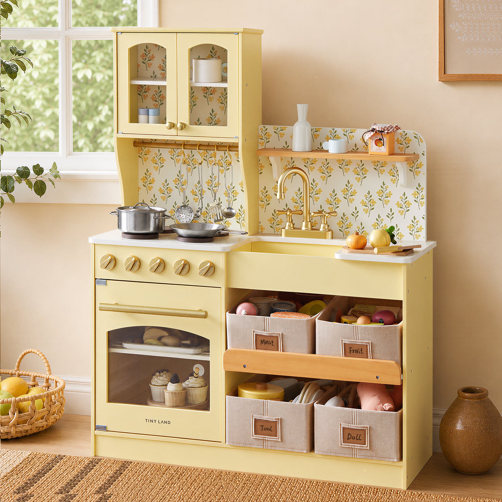 The Tiny Land® Wooden Play Kitchen with Storage for Creative Toddlers by Tiny Land, featuring multifunctional storage, play food, pretend cookware, and convenient baskets labeled Fruit, Vegetable, Tools, and Doll, sits against a wall adorned with a floral backsplash and a sunlit window—creating a cozy and eco-friendly play space.|Pale Yellow