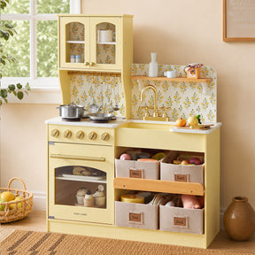 The Tiny Land® Wooden Play Kitchen with Storage for Creative Toddlers by Tiny Land, featuring multifunctional storage, play food, pretend cookware, and convenient baskets labeled Fruit, Vegetable, Tools, and Doll, sits against a wall adorned with a floral backsplash and a sunlit window—creating a cozy and eco-friendly play space.|Pale Yellow