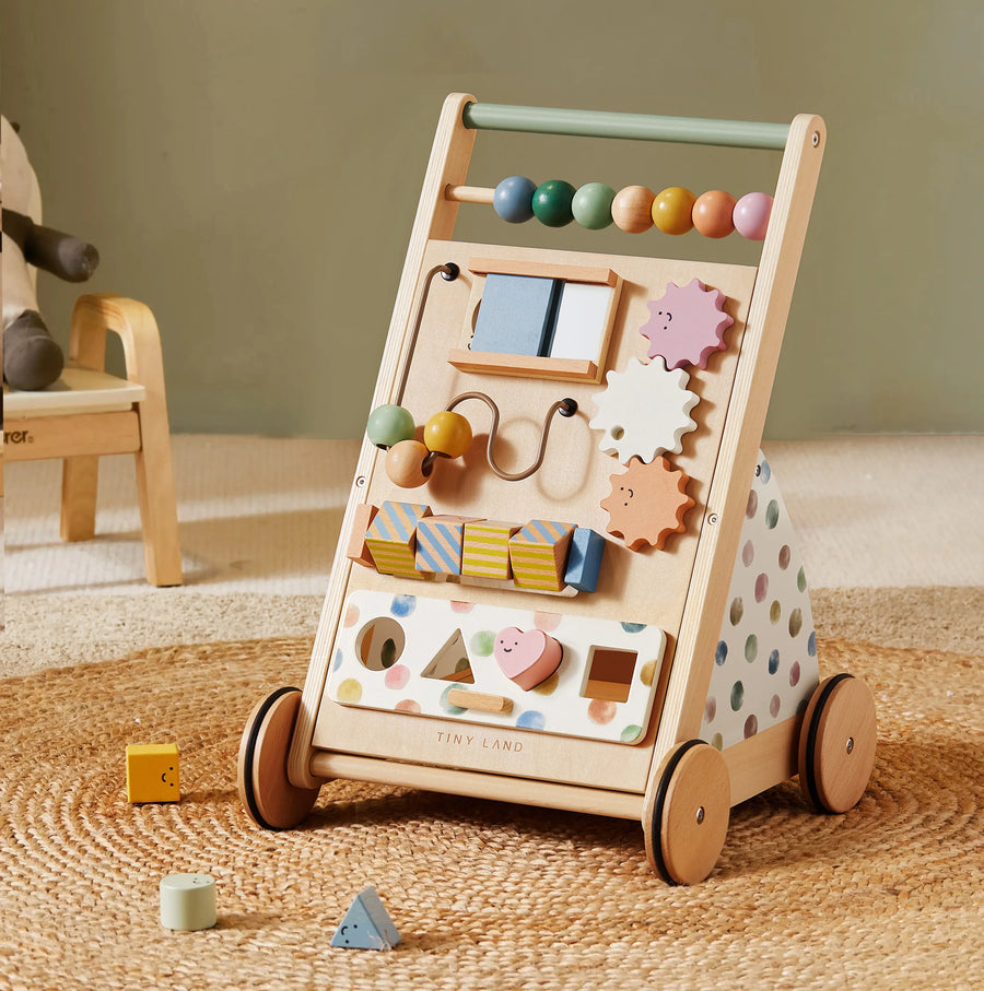The Tiny Land® Versatile Natural Wooden Baby Walker by Tiny Land is crafted from natural wood and features colorful beads, gears, and blocks in soft pastel shades. This baby learning walker is shown placed on a woven rug, accompanied by a small chair and scattered toy blocks nearby.