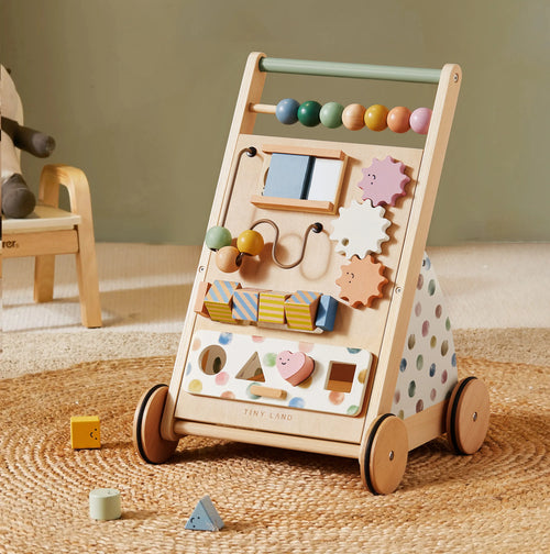 The Tiny Land® Versatile Natural Wooden Baby Walker by Tiny Land is crafted from natural wood and features colorful beads, gears, and blocks in soft pastel shades. This baby learning walker is shown placed on a woven rug, accompanied by a small chair and scattered toy blocks nearby.