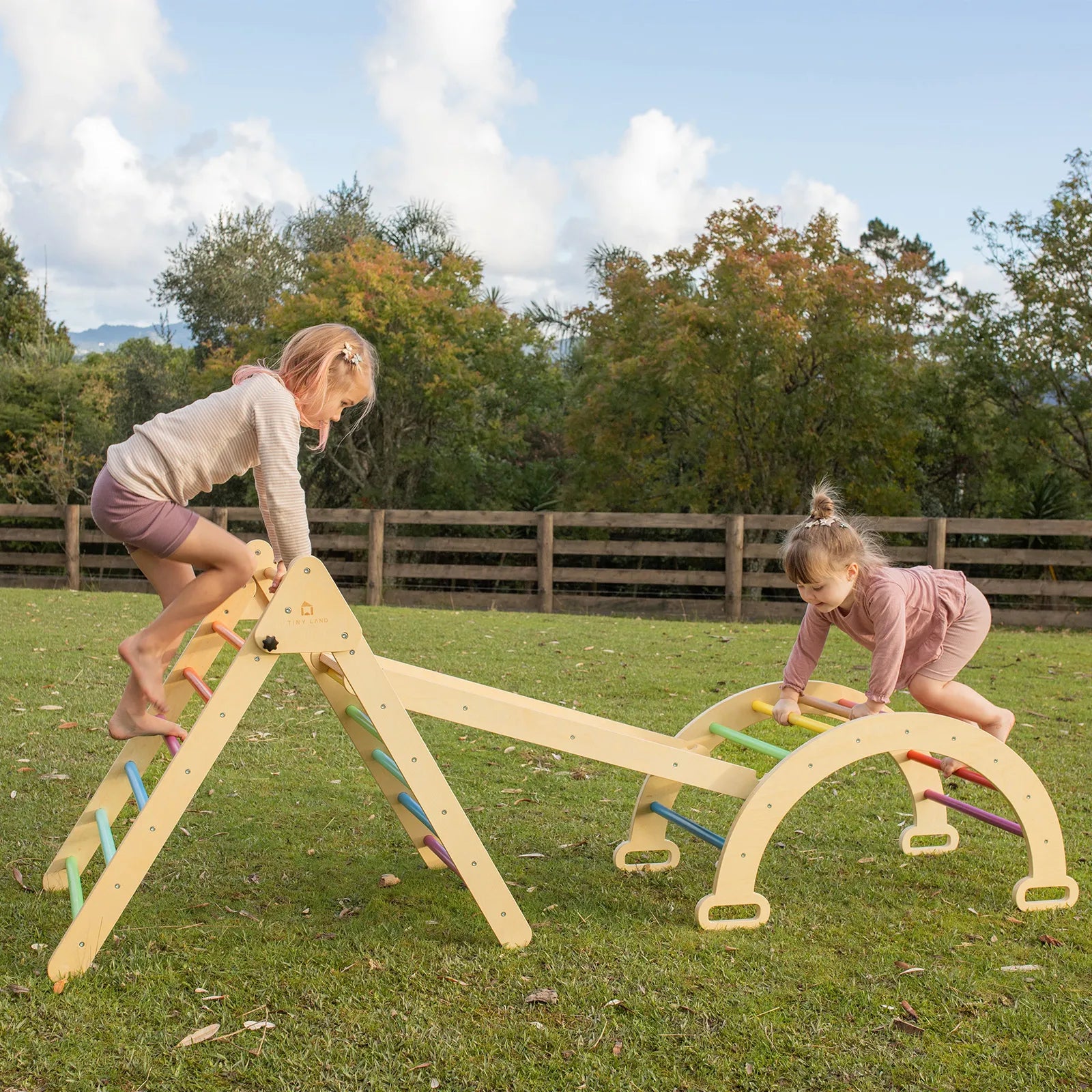 Two young children play on the Tiny Land® Pikler Triangle 7-in-1 Montessori Climbing Set by Tiny Land in a grassy outdoor area, surrounded by trees and a wooden fence under a partly cloudy sky.