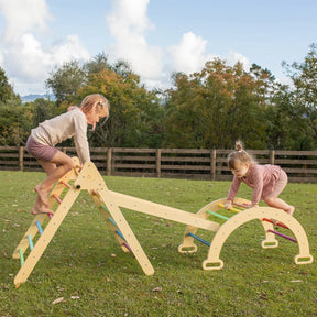 Two young children play on the Tiny Land® Pikler Triangle 7-in-1 Montessori Climbing Set by Tiny Land in a grassy outdoor area, surrounded by trees and a wooden fence under a partly cloudy sky.