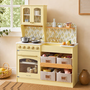 A Tiny Land® Wooden Play Kitchen with Storage for Creative Toddlers by Tiny Land, featuring a yellow finish, stove, oven, sink, shelves, and multifunctional storage bins, stands against a wall adorned with floral wallpaper. The kitchen is filled with pretend cookware, dishes, and play food, while a basket of plush food sits on the floor nearby.|Pale Yellow