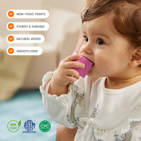 A baby in white bites the Tiny Land® Cottage Activity Cube - Rainbow by Tiny Land. The purple block features non-toxic paint, sturdy natural wood, smooth edges, and durability—making it a safe, developmental toy with visible certifications.