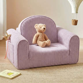 The Tiny Land® GentleSnug Kid Chair in light purple corduroy doubles as a sofa bed, shown with a beige teddy bear, star wand, and book in a cozy neutral-toned kids' room.