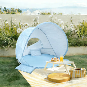 The Tiny Land® One-Stop Beach Toy & Tent Set in light blue gingham features a UV-protective tent with pillows, set on grass near a white table with snacks, plus a yellow cushion and striped mat nearby, all by a floral backdrop.