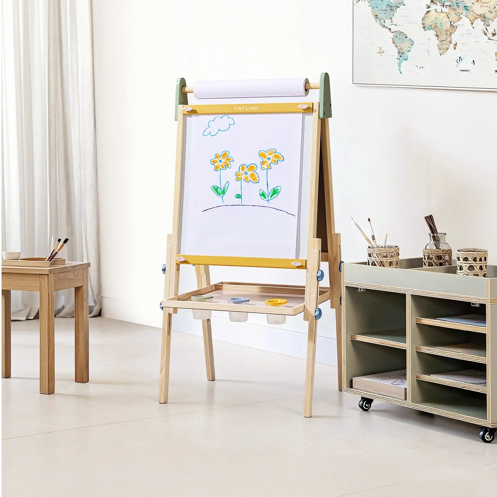 The Tiny Land® 3-in-1 Art Easel for Kids - Morandi stands in a bright room with a painting of yellow flowers and a cloud, next to art supplies, brushes on a small table, a storage cart, and a world map on the wall.
