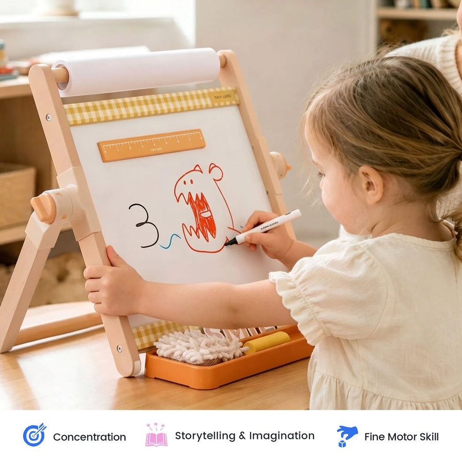 A young child uses the Tiny Land® Double-Sided Magnetic Tabletop Easel by Tiny Land to create a red monster with an open mouth, sitting at a light wooden table filled with art supplies in a cozy room. Icons below showcase the product’s benefits for concentration, imagination, and fine motor skills.|Oatmeal