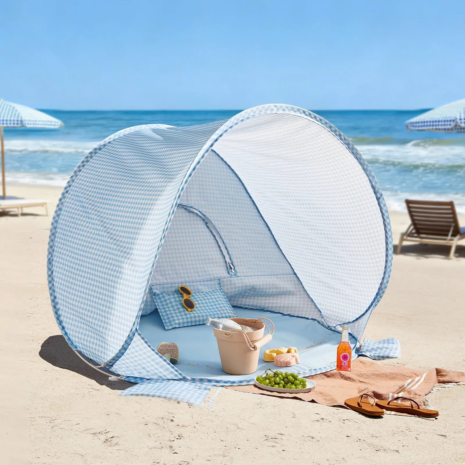 The Tiny Land® One-Stop Beach Toy & Tent Set in Light Blue Gingham sits on the sand near the ocean, with sunglasses and a beige bag inside. Nearby, a towel displays grapes, croissants, a drink, and sunscreen. Beach chairs and umbrellas are in the background.