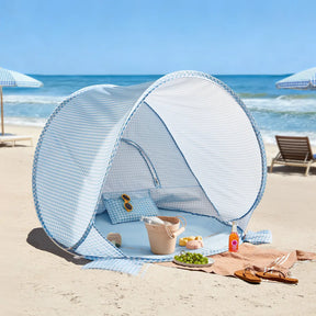 The Tiny Land® One-Stop Beach Toy & Tent Set in Light Blue Gingham sits on the sand near the ocean, with sunglasses and a beige bag inside. Nearby, a towel displays grapes, croissants, a drink, and sunscreen. Beach chairs and umbrellas are in the background.