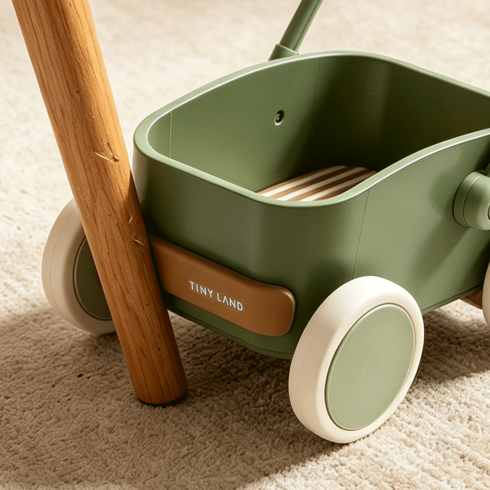 A close-up of the Test Tiny Land® JoyRide Baby Walker with Blocks - Sage, featuring white wheels and a wooden handle, rests on a beige carpet with sunlight casting soft shadows across the scene.