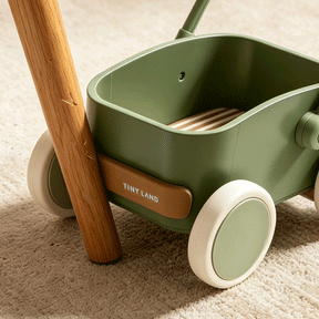 A close-up of the Test Tiny Land® JoyRide Baby Walker with Blocks - Sage, featuring white wheels and a wooden handle, rests on a beige carpet with sunlight casting soft shadows across the scene.