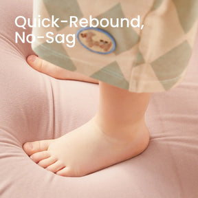 A child’s bare feet press into the soft, light pink cushion of the Tiny Land® GentleSnug Kid Chair by Tiny Land, crafted with high-density foam for comfort. The words “Quick-Rebound, No-Sag” appear in the upper left, while the child wears patterned pants adorned with a small animal patch.|Pink