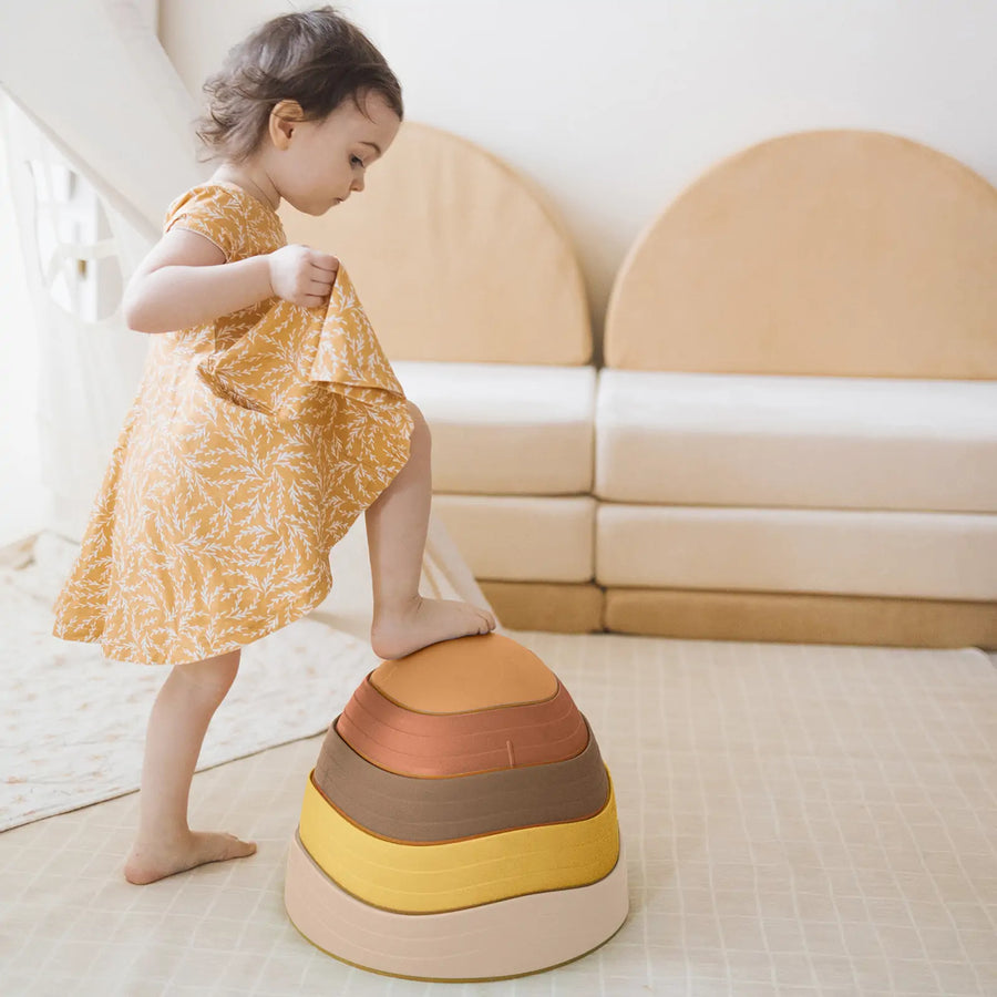 A young child in a yellow dress steps onto a stack of Tiny Land® Stepping Stones - Sunset Hues, building balance and coordination in a playroom with soft, neutral-colored furniture in the background.|Sunset Hues