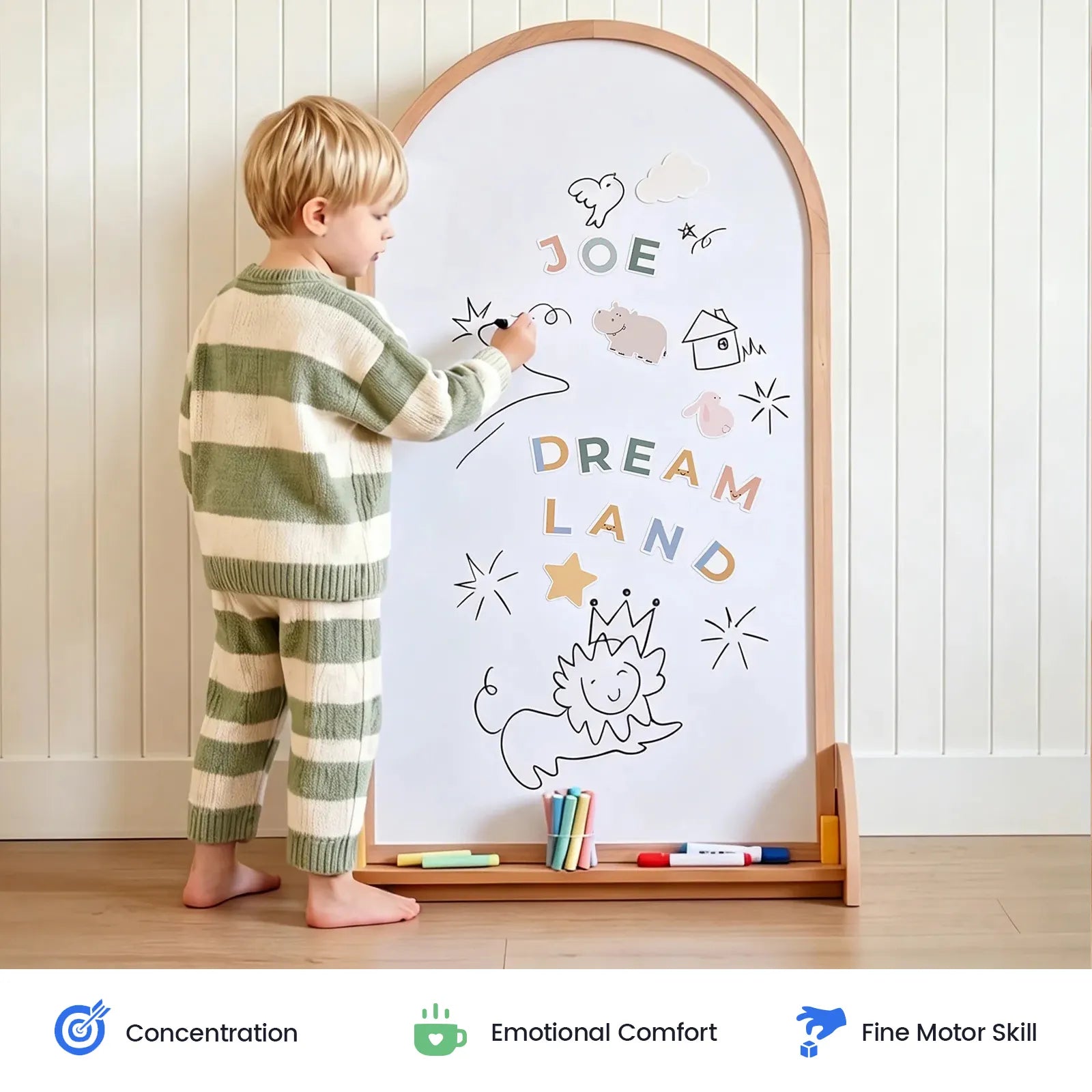 A young child in striped pajamas creates colorful doodles and writes “JOE DREAM LAND” on the Tiny Land® Double-Sided Arched Magnet Chalkboard, using vibrant markers. The playful scene showcases their concentration, emotional comfort, and fine motor skills, highlighted by icons below.