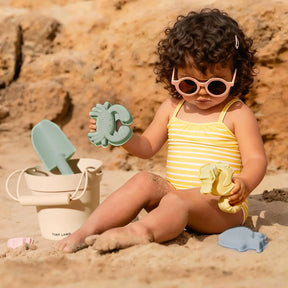 A toddler wearing a pastel yellow striped swimsuit and pink sunglasses plays on the sand with the Tiny Land® One-Stop Beach Toy & Tent Set, featuring a beige bucket and green shovel, with rocks in the background.