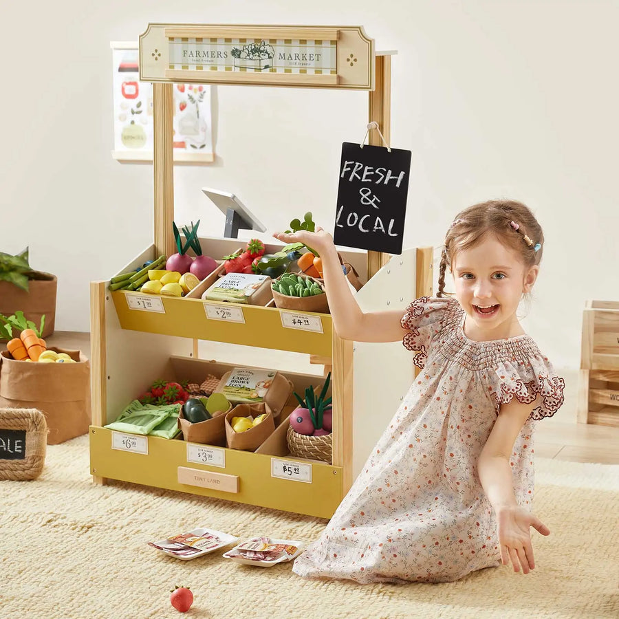A young girl in a floral dress smiles beside the Tiny Land® Kid's Grocery Store Playset, her wooden farmers market stand stocked with toy fruits and veggies as she proudly holds a "FRESH & LOCAL" sign for an imaginative pretend play experience.