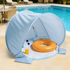The Tiny Land® One-Stop Beach Toy & Tent Set in light blue gingham shelters an orange swim ring, sunglasses, and a glass of orange juice on a white towel by the pool, with a wicker chair and table nearby.