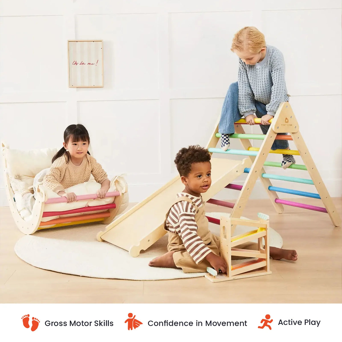 Three young children play indoors on the Tiny Land® 7-in-1 Rainbow Climbing Set with Play Pad, featuring a vibrant Pikler triangle, Montessori-inspired climber, slide, and rocker. The bright, minimal room is accented by a round rug. Icons below showcase benefits like gross motor skill development, building confidence, and encouraging active play.