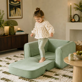 A young child smiles while standing on the Tiny Land® GentleSnug Kids Sofa Chair in a cozy living room decorated with candles, plants, and framed art.