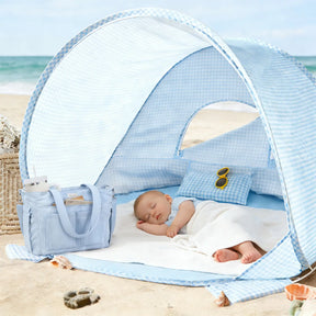 A baby naps under the Tiny Land® One-Stop Beach Toy & Tent Set in light blue gingham on the sand, with a matching diaper bag, tiny sunglasses, and sand toys nearby. The ocean and sky provide a serene backdrop.
