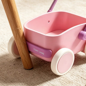 A close-up of the Tiny Land® 3-in-1 Wooden Baby Walker Wagon - Pink, featuring white wheels, is shown next to a wooden table leg on a beige carpet.