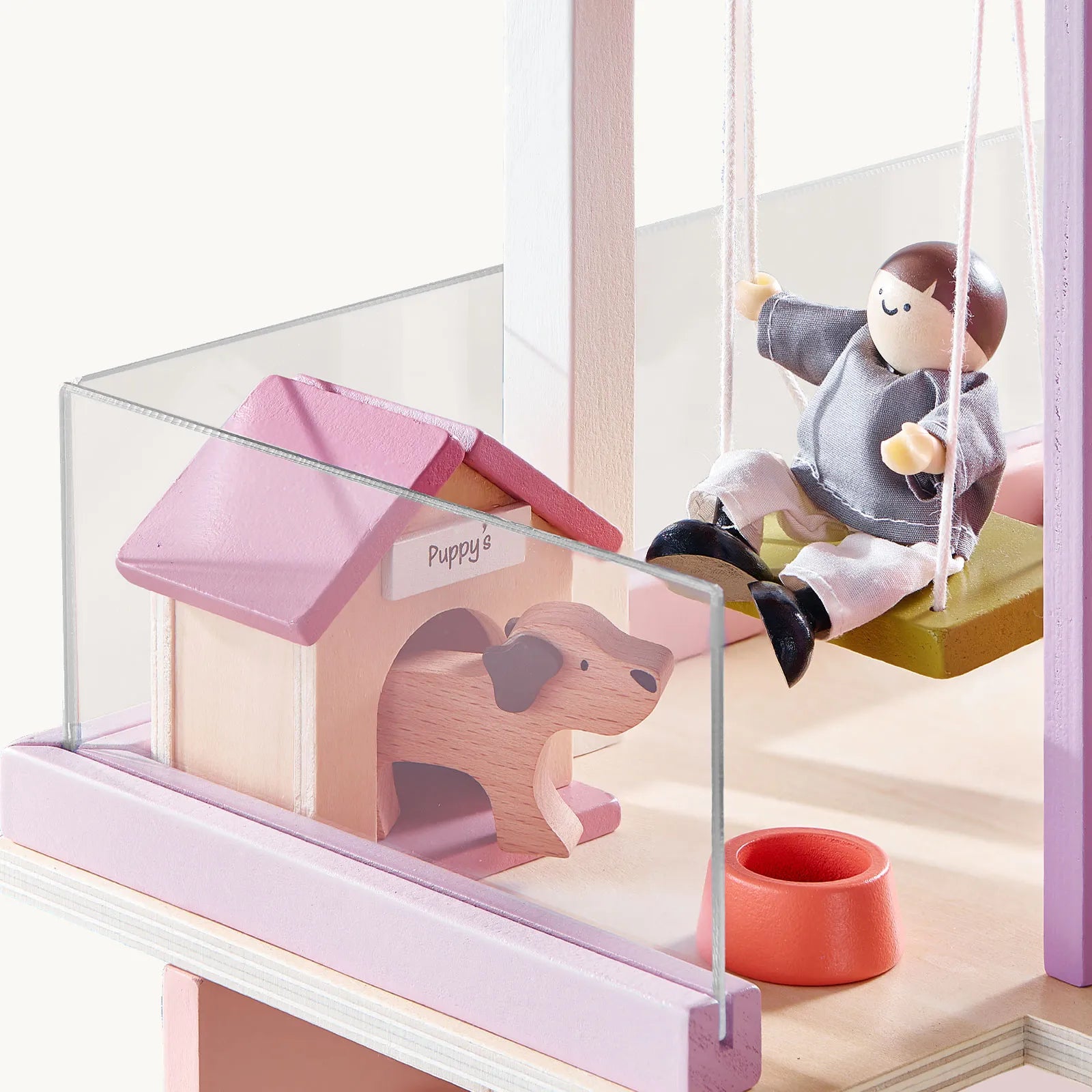 A Tiny Land® Sweetwood Cottage Dollhouse features a wooden doll on a swing next to a pink and beige doghouse labeled "Puppy," with a dog cutout entrance, red food bowl, and enclosed by a clear plastic fence.|Pink
