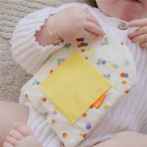 A baby in white holds a soft, dotted cloth with a yellow patch from the Tiny Land® Montessori Early Skills Baby Set (7–12 months), perfect for sensory exploration and fine-motor development by Tiny Land.
