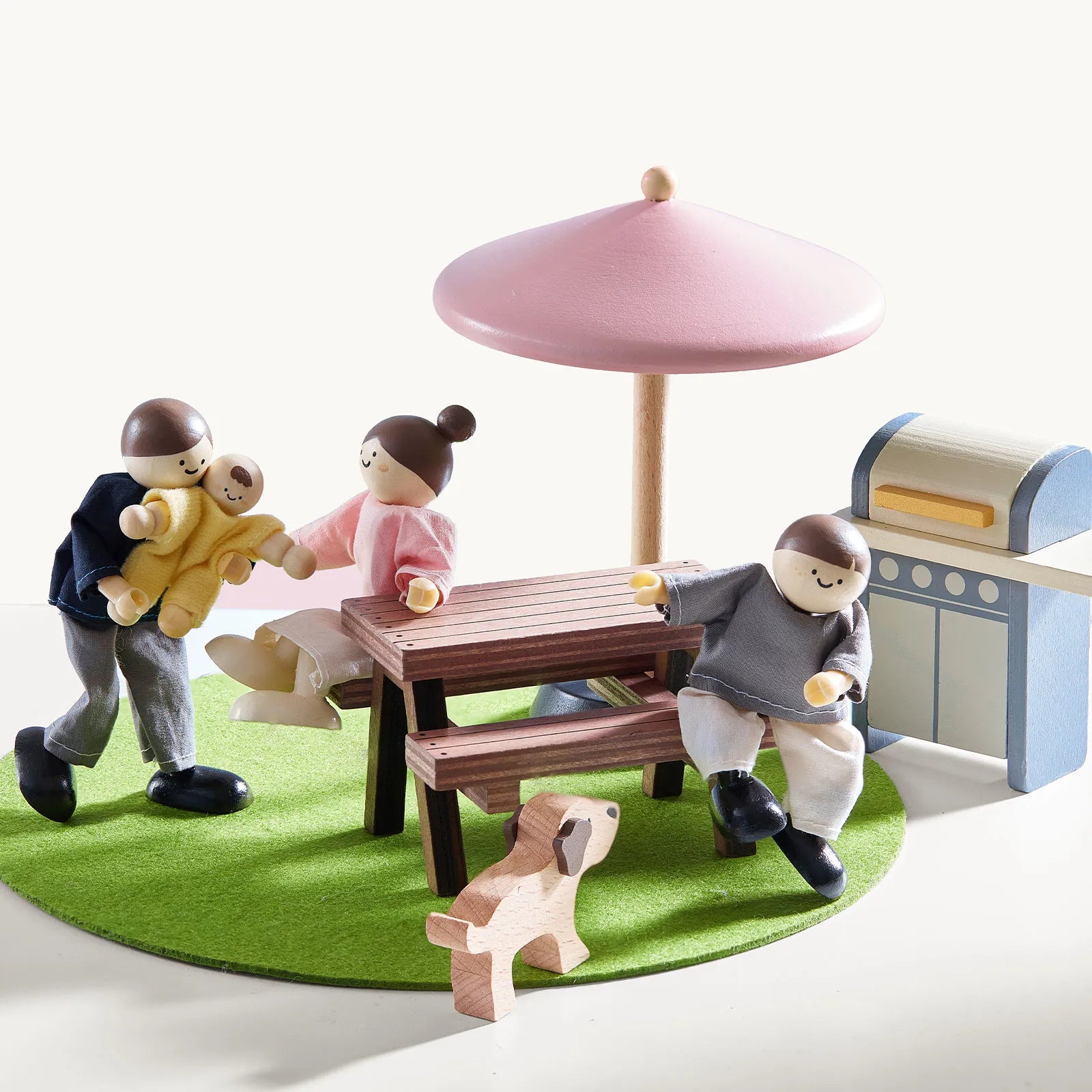 In the Tiny Land® Sweetwood Cottage Dollhouse by Tiny Land, three wooden dolls and a baby gather around a picnic table with a pink umbrella, with a grill and dog figure nearby—perfect for imaginative outdoor family barbecues.|Pink