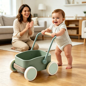 A smiling baby pushes the Test Tiny Land® JoyRide Baby Walker with Blocks - Sage, featuring adjustable speed control, in a bright living room as a woman behind claps and smiles encouragingly.