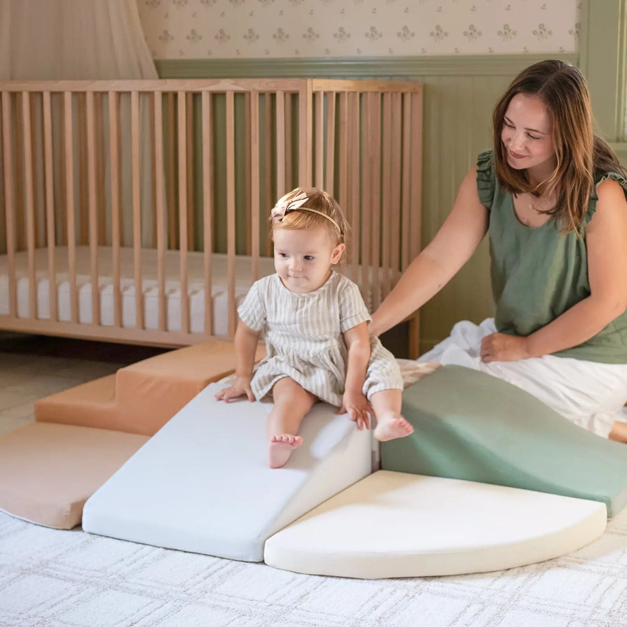 A toddler explores sensory play on the Tiny Land® PlayNest Climbing & Ball Pit Set - Blue, accompanied by a smiling woman in a nursery with a wooden crib and patterned wallpaper—a cozy play space from Tiny Land.