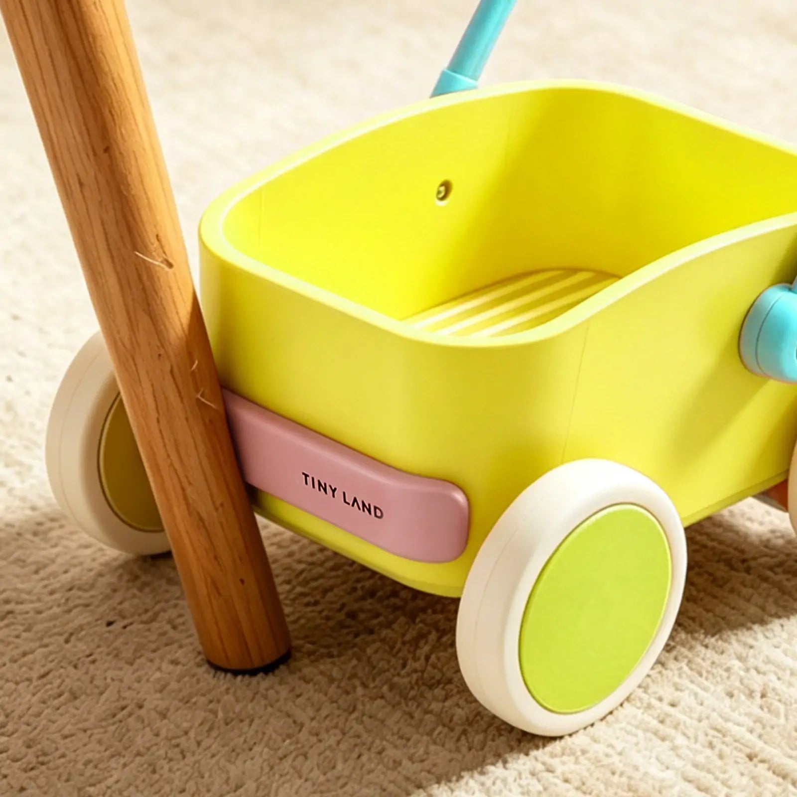 A close-up of the Tiny Land® JoyRide Baby Walker with Blocks - Spring Color by Test, styled like a toy wagon with white and green wheels, adjustable speed control, blue handle, and pink label, on a beige carpet next to wooden furniture.
