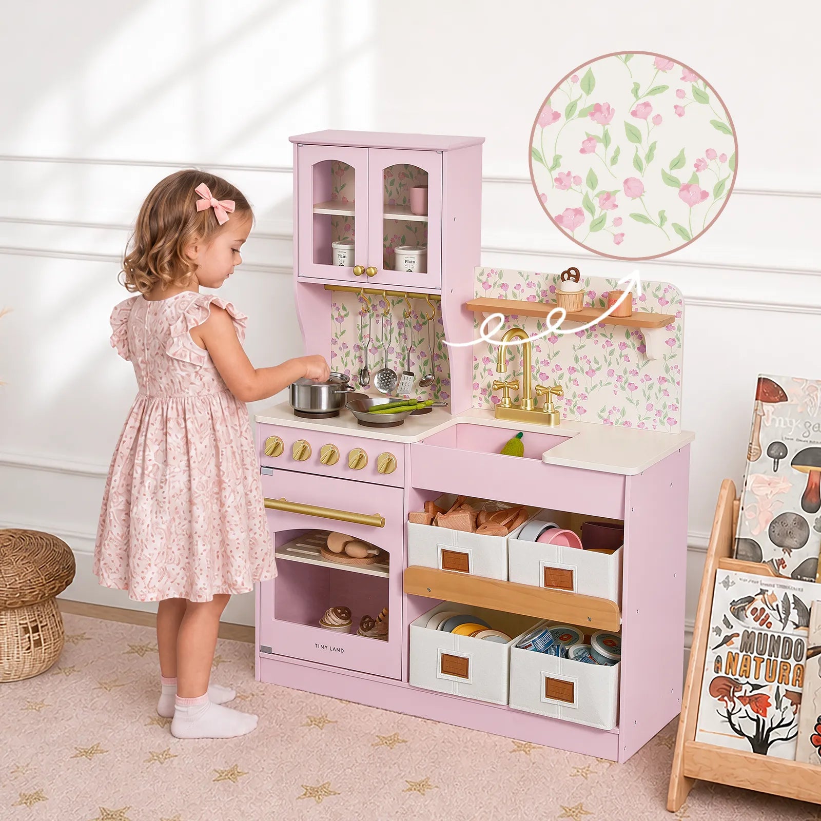 A young girl in a pink dress plays with the Tiny Land® Wooden Play Kitchen with Storage for Creative Toddlers by Tiny Land, featuring pretend food, utensils, and realistic cookware, in a cozy room accented with floral wallpaper.|Sakura Pink