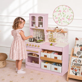 A young girl in a pink dress plays with the Tiny Land® Wooden Play Kitchen with Storage for Creative Toddlers by Tiny Land, featuring pretend food, utensils, and realistic cookware, in a cozy room accented with floral wallpaper.|Sakura Pink