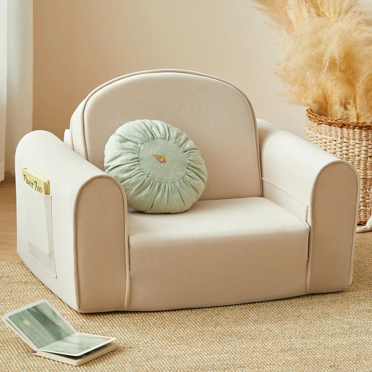 The Tiny Land® GentleSnug Kid Chair by Tiny Land, a small beige armchair featuring a round green cushion and crafted with high-density foam for lasting comfort, sits atop a tan rug. A book rests in the chair’s side pocket, while a woven basket filled with pampas grass is visible in the background.|Beige