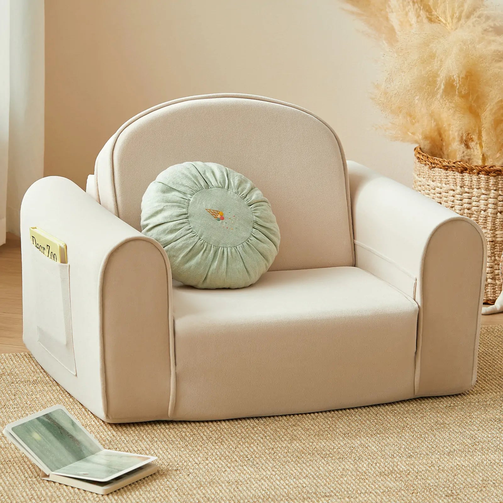 The Tiny Land® GentleSnug Kid Chair by Tiny Land, a small beige armchair featuring a round green cushion and crafted with high-density foam for lasting comfort, sits atop a tan rug. A book rests in the chair’s side pocket, while a woven basket filled with pampas grass is visible in the background.|Beige