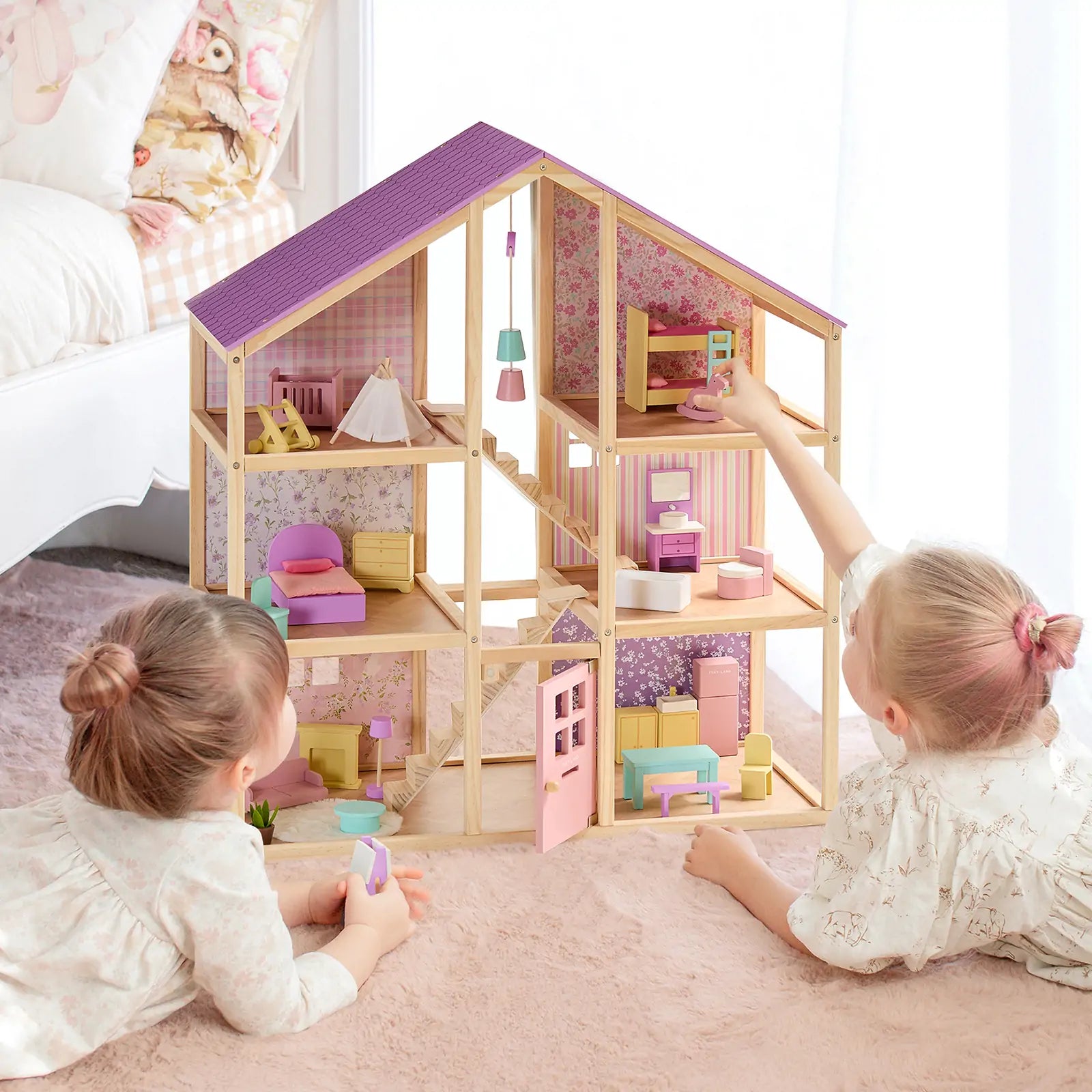 Two young children play with the Tiny Land® Sweetwood Unicorn Dollhouse, complete with pastel-colored rooms and Tiny Land wooden poseable dolls, on a pink rug in a bright bedroom.