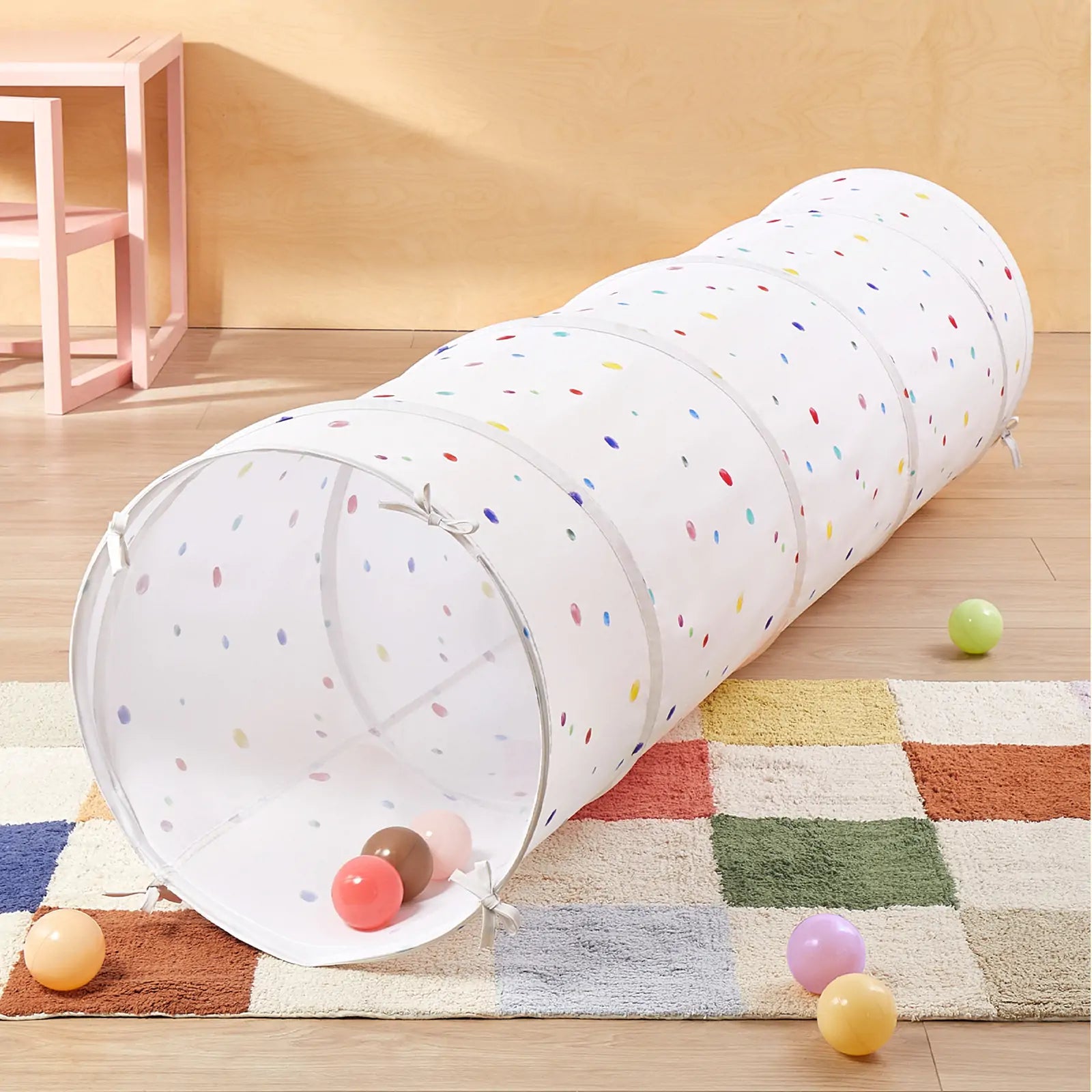The Tiny Land® Adventure Play Tunnel - Colorful Polka Dots by Tiny Land lies on a checkered rug with colorful plastic balls inside and around it. A pink table and chair are in the background against a light wood wall.|white