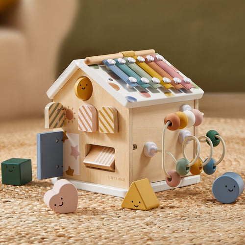 The Tiny Land Cozy Learning Playset—Playmat + Activity Cube—features pastel xylophone bars, shape sorters, a bead maze, and geometric blocks on a woven rug, making it an ideal developmental toy for toddlers.