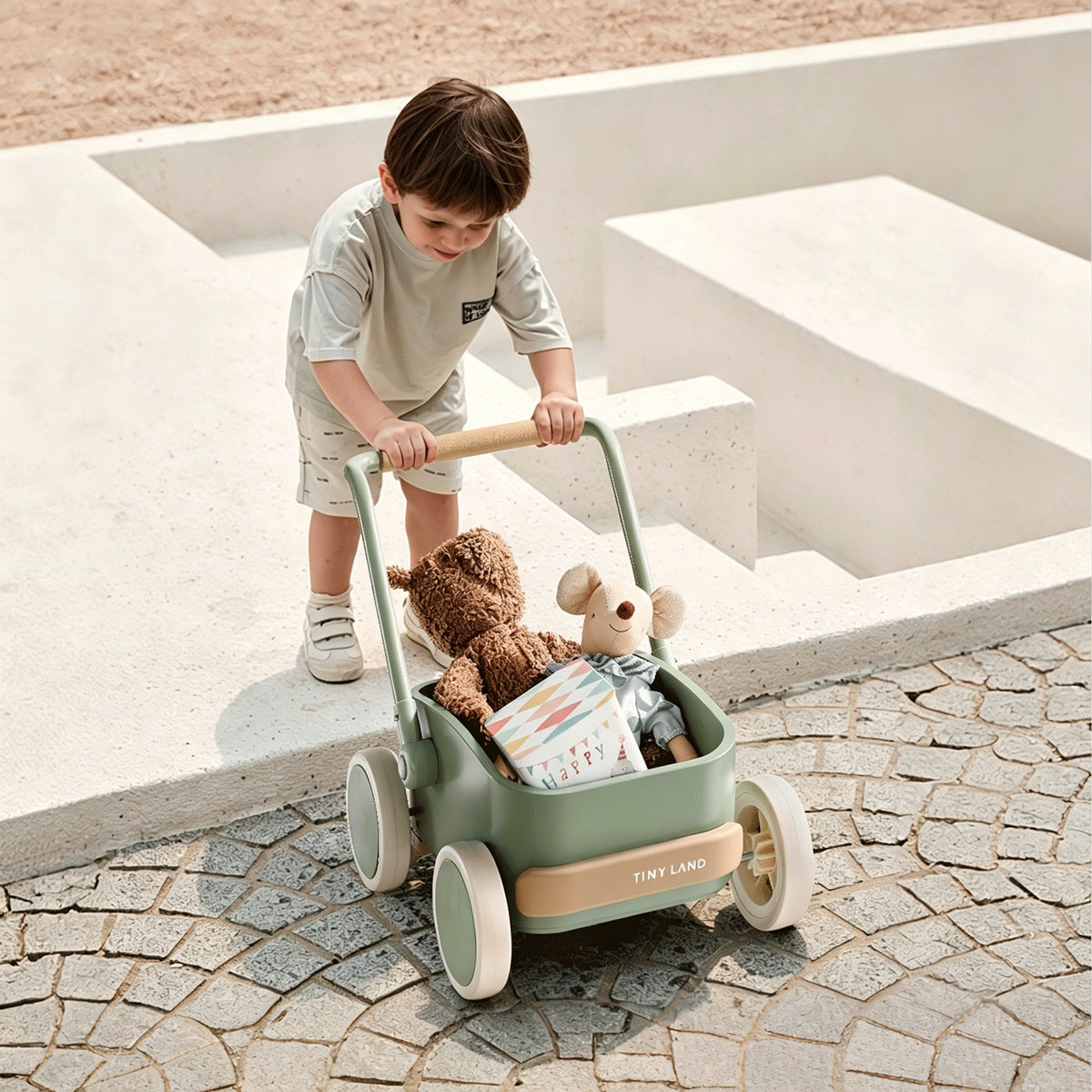 A young child pushes the Tiny Land® JoyRide Baby Walker with Blocks - Sage by Test outdoors, carrying a teddy bear, gray stuffed animal, and colorful book near stone steps and a paved walkway.