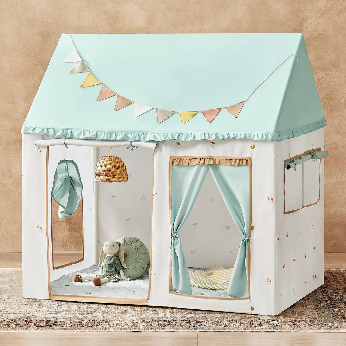 The Tiny Land® Sweet Woodland Kids Play Tent by Tiny Land INC features a pastel indoor design with a blue roof, pennant banner, hanging basket light, curtains, cushion, stuffed rabbit, green pillow, and gray patterned play mat against a tan wall.