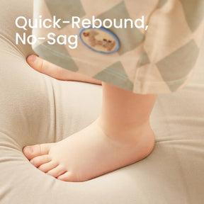 A close-up of a child's bare feet on the plush surface of the Tiny Land® GentleSnug Kid Chair - Beige White. The text "Quick-Rebound, No-Sag" appears in the top left. The child wears light-colored, patterned pants. Brand: Tiny Land.