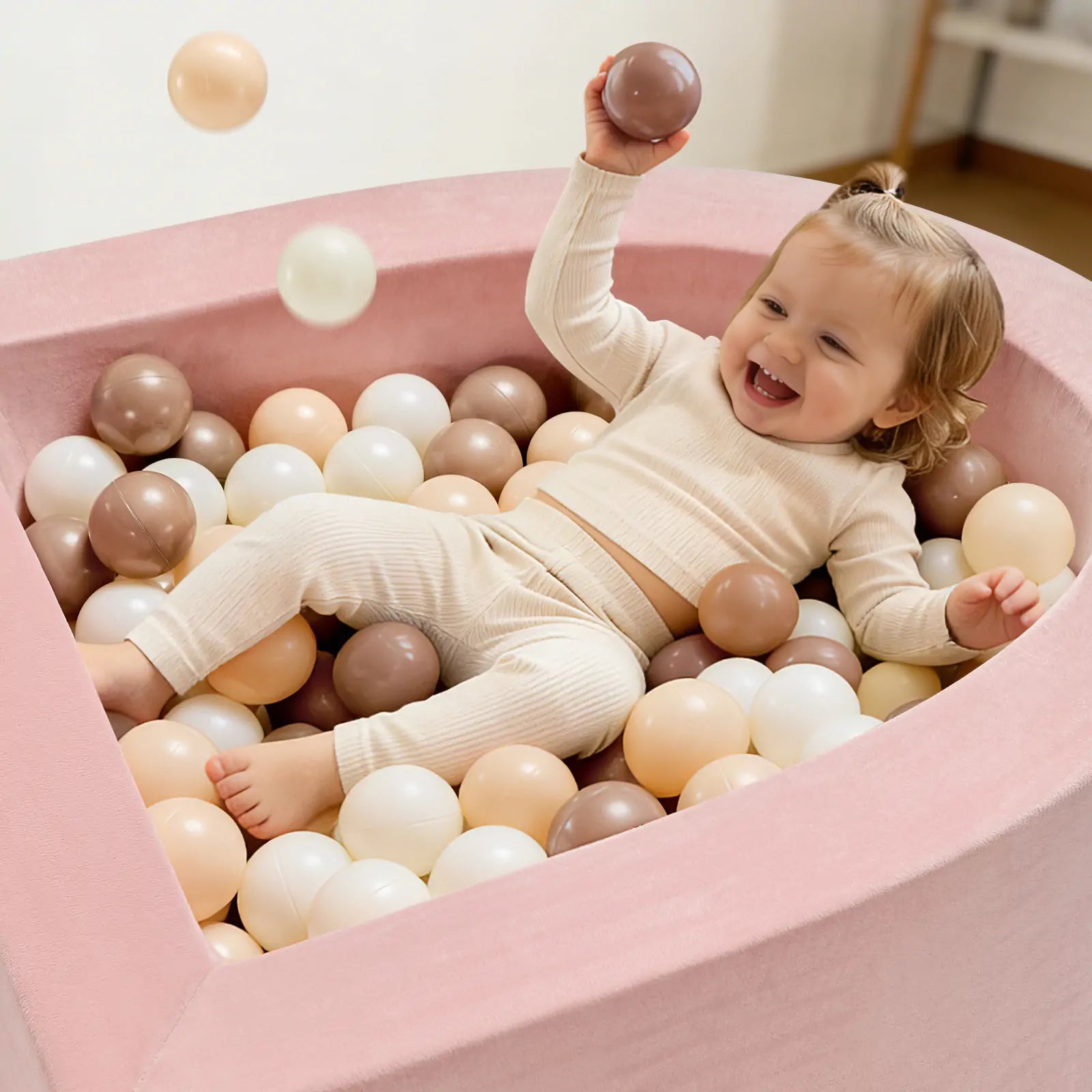A smiling toddler in cream clothes joyfully plays in the Tiny Land® PlayNest Ball Pit by Tiny Land, filled with 200 white, beige, and brown ocean balls, holding one up while enjoying the soft pink foam play space.|Pink