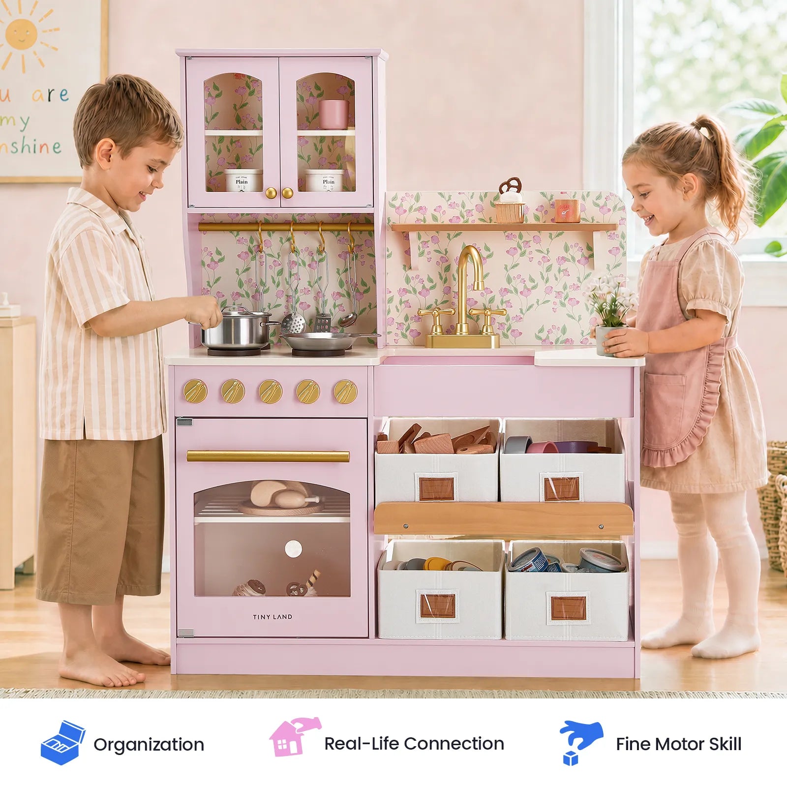 Two young children play at the Tiny Land® Wooden Play Kitchen with Storage for Creative Toddlers, pretending to cook with eco-friendly pretend cookware, pots, and utensils. This pink wooden kitchen from Tiny Land features shelves with baskets and toy food, spacious cabinets, and a sink. Icons below highlight skills such as organization and fine motor development.|Sakura Pink
