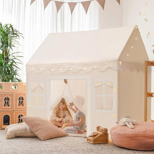 Two children enjoy the Tiny Land Bright Play & Art Set, featuring a colorful easel and a cream playhouse with lights, surrounded by pillows and toys in a bright, plant-filled room—perfect for imaginative indoor or outdoor fun|white